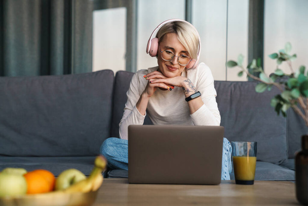 Smiling mid aged blonde woman in headphones studying online via laptop computer in the living room - Photo, Image