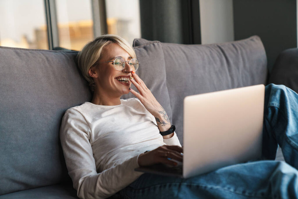 Smiling laughing mid aged blonde woman studying online via laptop computer in the living room - Photo, Image