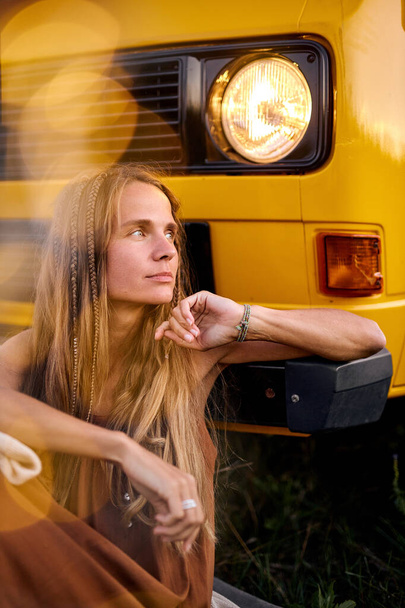beautiful woman is looking at side, posing, sitting on ground next to yellow van - Photo, Image
