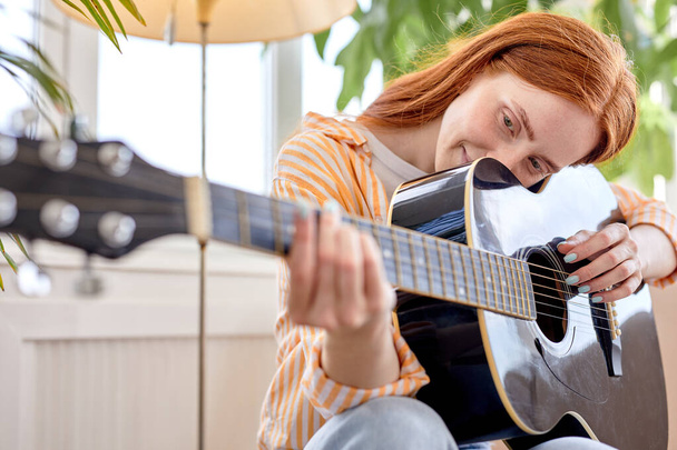 Portret van jonge roodharige vrouw in casual dragen spelen gitaar, Thuis - Foto, afbeelding
