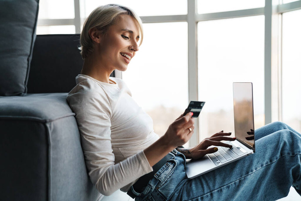 Smiling blonde mid aged woman shopping online with credit card and laptop computer on a couch at home - Photo, image