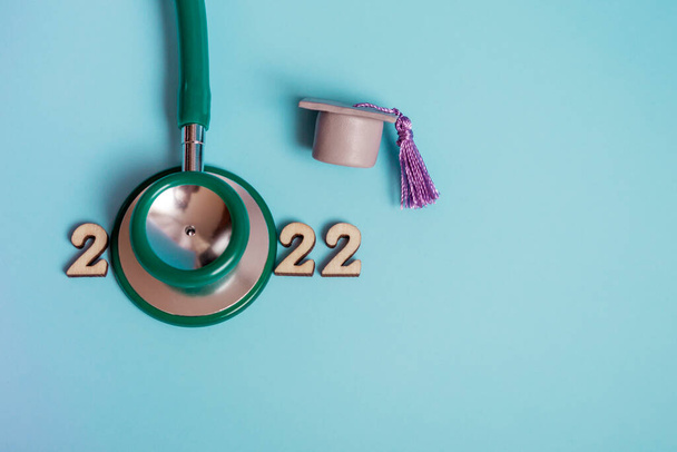 Absolventenmütze mit Stethoskop und Zahlen 2022 auf blauem Hintergrund, Nahaufnahme mit Kopierraum. Medizinisches Ausbildungskonzept. - Foto, Bild