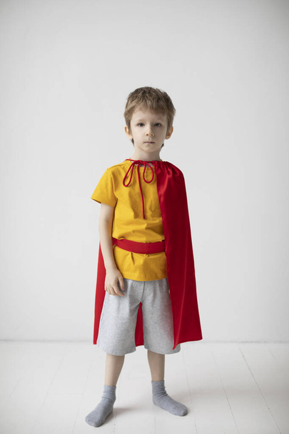 Portrait of a serious little boy dressed like superhero standing alongside the white wall, having fun at home. - Фото, изображение