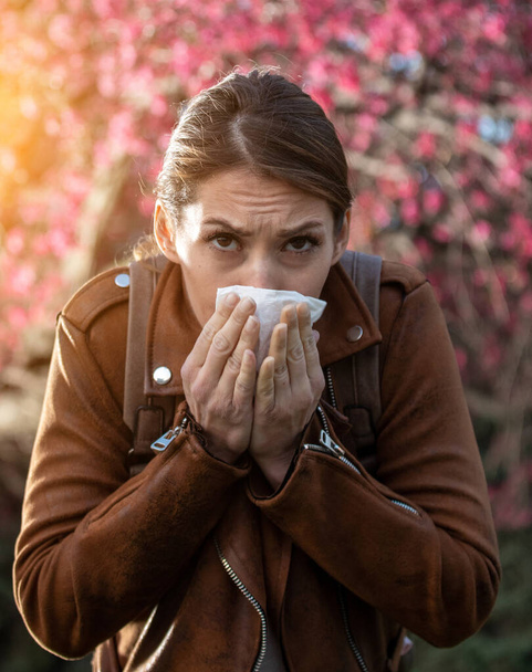 Young pretty girl blowing nose in front of blooming tree. Spring allergy reaction - Photo, Image