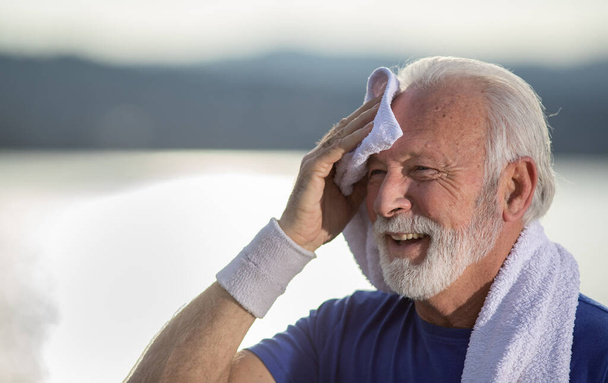 Улыбающийся пожилой мужчина с седыми волосами и бородой отдыхает после тренировки, вытирает лоб полотенцем, на открытом воздухе - Фото, изображение