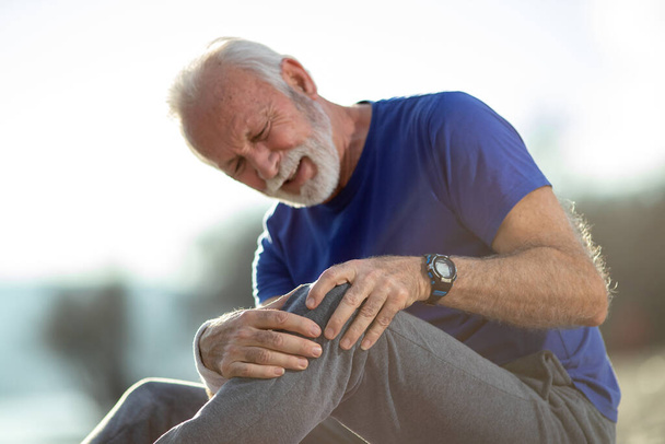 Senior man zit in sportkleding voelt pijn in zijn knie na het sporten in de natuur selectieve focus - Foto, afbeelding