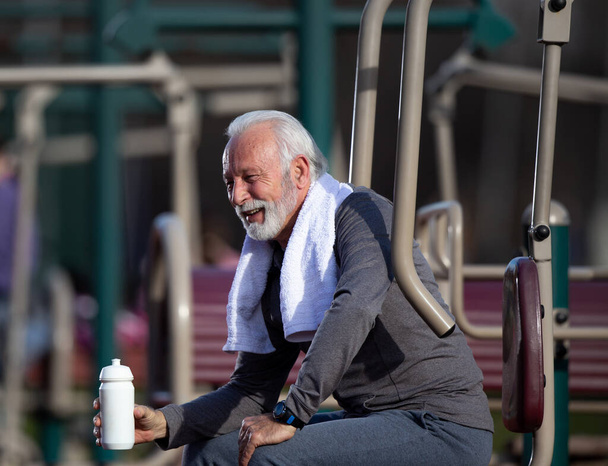 Uomini anziani che bevono acqua dopo aver usato attrezzature nella palestra pubblica all'aperto e indossando un asciugamano intorno al collo - Foto, immagini