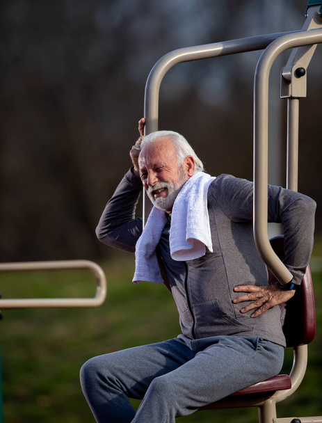 Oudere man met baard die pijn in de onderrug voelt na inspanning in een openbare fitnessruimte buiten - Foto, afbeelding