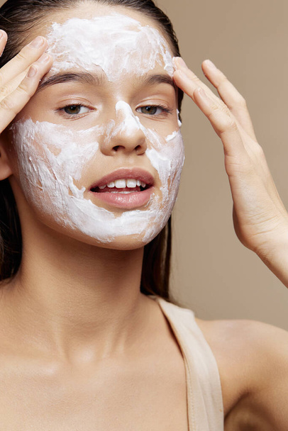 sorridente giovane donna crema sulla guancia posa cosmetici per pulire la pelle sfondo beige - Foto, immagini
