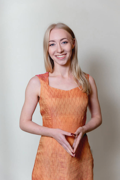 красивая улыбающаяся блондинка пристально смотрит на камеру и жесты руками, одетыми в открытое оранжевое платье - Фото, изображение