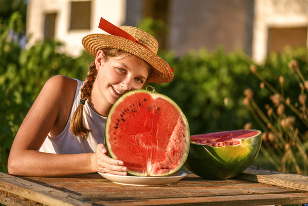 Porträt eines jungen glücklichen Mädchens mit Hut und der Hälfte der roten frischen Wassermelone, die es sich im Freien gemütlich macht. Teenager genießen das Sommerleben. Sommerliches Lifestyle-Konzept. Glück, Freude, Feiertag Independence Day - Foto, Bild