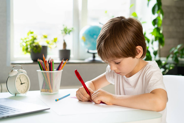 Маленький мальчик делает уроки дома за столом перед ноутбуком, дистанционное обучение онлайн образования. - Фото, изображение