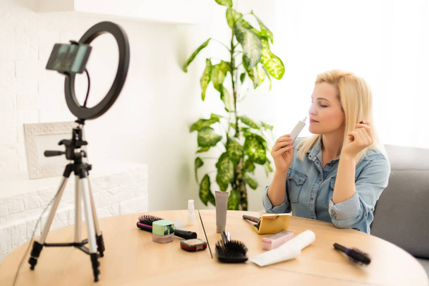 blogger woman recording video on smartphone and lighting herself with ring lamp at table in light room - Photo, Image