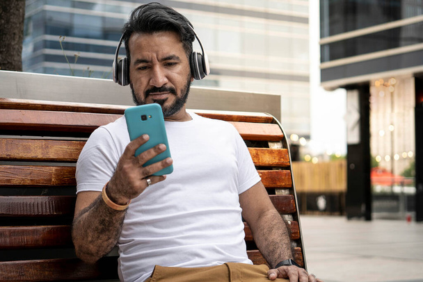 homem latino com barba sentado em um banco olhando para seu telefone celular em um ambiente urbano - Foto, Imagem