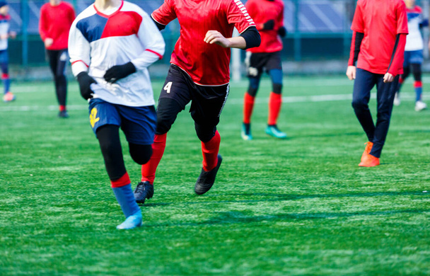 Chłopak w czerwonej białej odzieży sportowej gra w piłkę na boisku, drybluje piłkę. Młodzi piłkarze nastolatki z piłką na zielonej trawie. - Zdjęcie, obraz