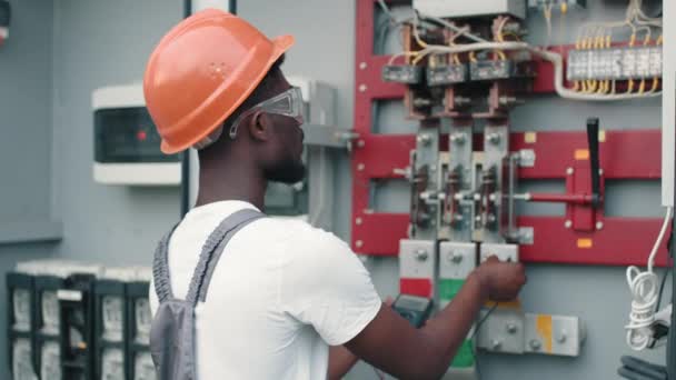 African american man in grey overalls, safety helmet and glasses smiling and looking at camera while standing near switchgear at solar station. Production of alternative energy. - Footage, Video