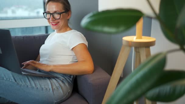 Smiling woman with glasses is sitting on the couch and working on a laptop or chatting with someone. Concept of remote work. - Footage, Video