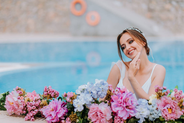 Beautiful bride in a beautiful white swimsuit and garlands of flowers, standing in swimming pool with flower on turquoise water, beautiful girl in water pool in sunny day - Foto, afbeelding