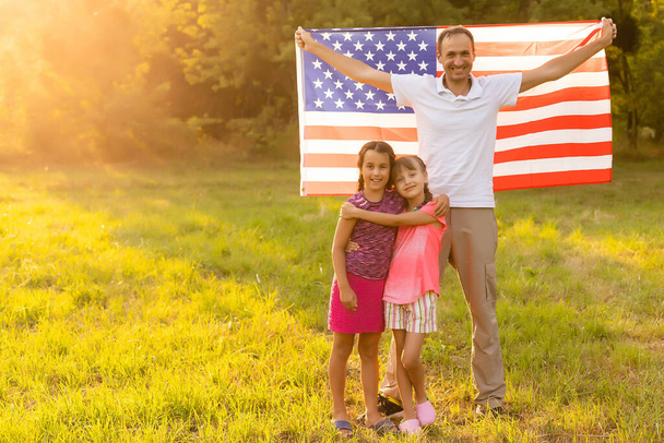 Ευτυχισμένη οικογένεια με αμερικανική σημαία σε χωράφι με σιτάρι το ηλιοβασίλεμα. Ημέρα Ανεξαρτησίας, 4 Ιουλίου. - Φωτογραφία, εικόνα