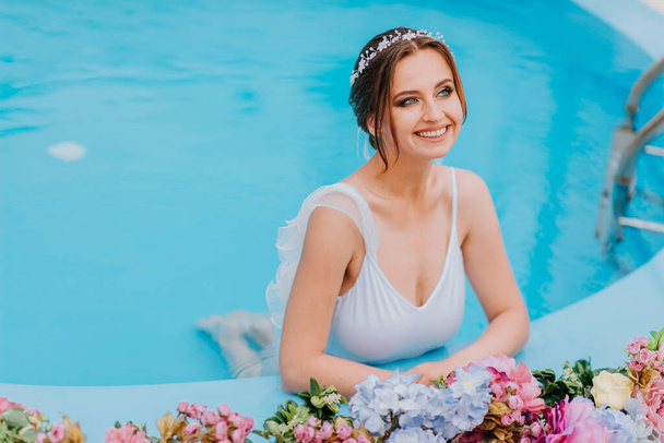 Beautiful bride in a beautiful white swimsuit and garlands of flowers, standing in swimming pool with flower on turquoise water, beautiful girl in water pool in sunny day - Фото, изображение