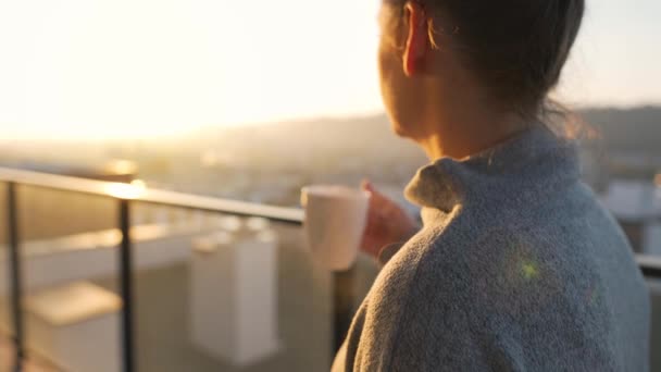 Kobieta zaczyna dzień od filiżanki herbaty lub kawy na balkonie o świcie, w zwolnionym tempie. Krajobraz miasta w tle - Materiał filmowy, wideo