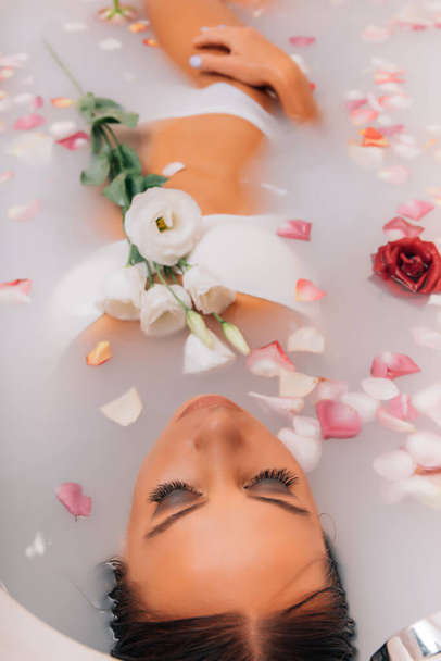 Luxus spa nő. elegáns szemek csillogás lány selyem bőr szórakozás feküdt vízfürdőben pihentető között rózsaszirmok. A nő egy tejjel teli wellness fürdőt vesz. - Fotó, kép