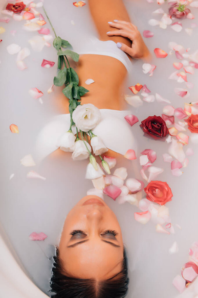 Luxus spa nő. elegáns szemek csillogás lány selyem bőr szórakozás feküdt vízfürdőben pihentető között rózsaszirmok. A nő egy tejjel teli wellness fürdőt vesz. - Fotó, kép