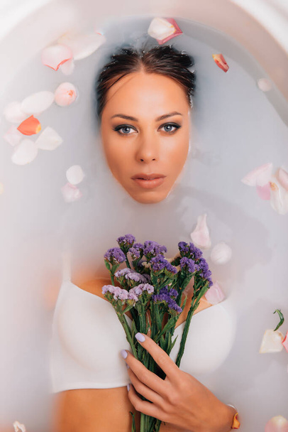 luxury spa woman. elegant eyes glamour girl with silk skin having fun laying in water bath relaxing among rose petals. The woman takes a wellness bath filled with milk. - Foto, Bild