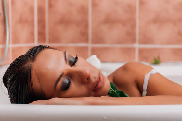 luxe spa vrouw. elegante ogen glamour meisje met zijden huid plezier te leggen in het water bad ontspannen tussen rozenblaadjes. De vrouw neemt een wellnessbad gevuld met melk. - Foto, afbeelding