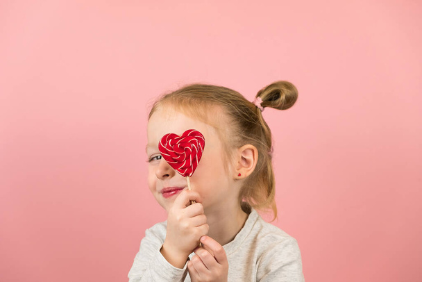 Funny little blonde girl smiling and playing with red heart shape lollipop caramel on pink background. Valentines Day or Mothers Day concept - 写真・画像
