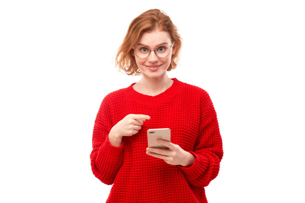 Shocked joyful redhead girl points finger at smartphone blank screen in spectacles and red clothes isolated on white studio background - Fotoğraf, Görsel