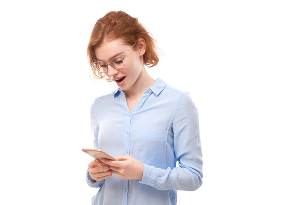 Happy smiling redhead girl holding smartphone in hands in spectacles and business shirt isolated on white studio background, received a joyful message - Photo, image