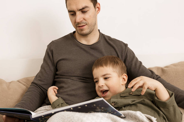 Vader en zoon lezen thuis een boek op het bed. Jonge aantrekkelijke man en kleine jongen rustend in de slaapkamer. Natuurlijke aardekleuren. Gezellige omgeving. Vader leest een sprookje voor zijn kind. - Foto, afbeelding