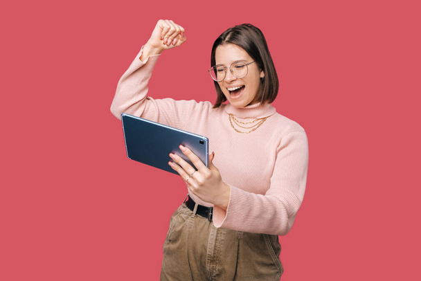 Mladá veselá trendy žena drží tablet a slaví přes růžové pozadí - Fotografie, Obrázek