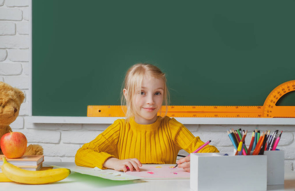 Back to school. Cute pupil girl drawing at the desk. Child in the class room with blackboard on background. Education, learning and children concept. - Fotó, kép