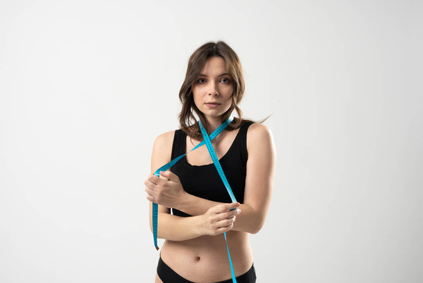 Portrait of a confused young brunette woman in a black underwear holding measuring tape around her neck over white background. Weight loss, slim body, healthy lifestyle concept. - Foto, afbeelding