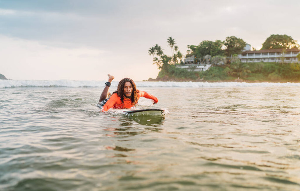 Black long-haired man paddling on long surfboard to the surfing spot in Indian ocean. Palm grove litted sunset rays in the background. Extreme water sports and traveling to exotic countries concept. - 写真・画像