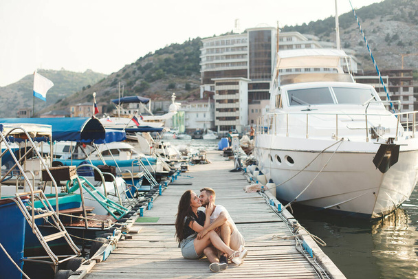 Lovers, guy and girl, are sitting on a wooden pier, holding hands and laughing - Фото, зображення