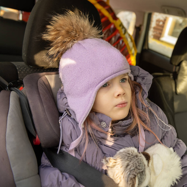 A girl of 5 years old is sitting with a soft toy in her hands in a car, fastened with a seat belt in a child's chair. - Photo, Image