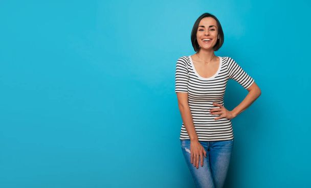 Close up photo of a beautiful smiling confident young brunette woman is posing in a striped t-shirt and isolated on blue background - Photo, image