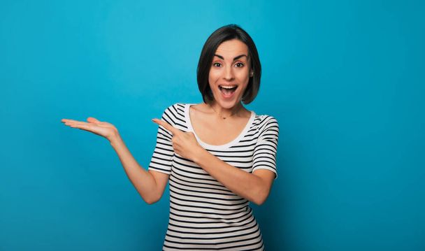 Photo of an excited attractive brunette woman in casual wear while she pointing at something advert on her arm and isolated on blue background - Φωτογραφία, εικόνα