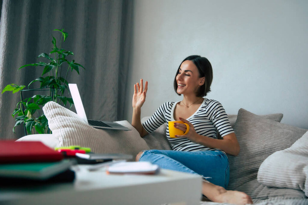 Cute smiling freelancer woman working on a laptop and has an online conversation while she sitting on the big couch and drinking coffee at home - Valokuva, kuva