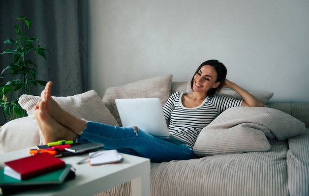 Young beautiful smiling woman with laptop while she lay on the big couch at home and relaxing - Foto, imagen