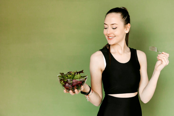 Nuori viehättävä nainen mustissa urheiluvaatteissa, poninhäntä, syö salaattia, vihreällä taustalla. Terveellinen elämäntapa käsite - Valokuva, kuva