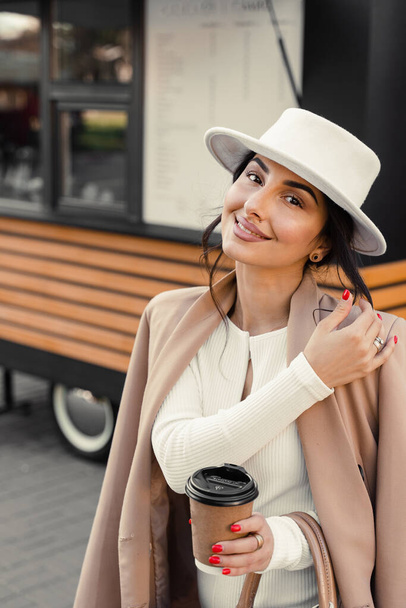 Model z filiżanką kawy w pobliżu kawiarni. Dziewczyna nosiła białą sukienkę, kurtkę i kapelusz pozowanie w pobliżu kawiarni - Zdjęcie, obraz