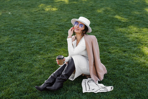 Μόδα μοντέλο με φλιτζάνι καφέ κάθισμα στο γρασίδι. Ελκυστική γυναίκα φορούσε καπέλο, φόρεμα, σακάκι και γυαλιά ηλίου - Φωτογραφία, εικόνα