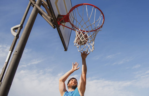 Basketball player. Sports and basketball. A young man jumps and throws a ball into the basket. Blue sky and court in the background. - Φωτογραφία, εικόνα