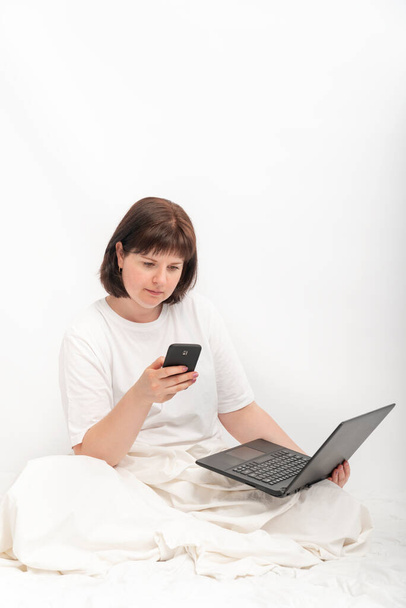 Business woman with laptop and phone in bed against a white wall. Young woman works sitting in bed. Vertical frame. - Photo, Image