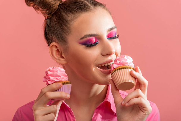 Chica joven en camisa rosa sobre fondo rosa muerde magdalena. Maquillaje juvenil profesional en tonos rosados - Foto, Imagen