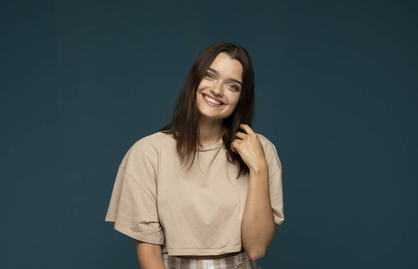 Close-up portrait of young brunette emotional woman in beige t-shirt with a big smile. Beautiful model posing in studio over blue background. - Φωτογραφία, εικόνα